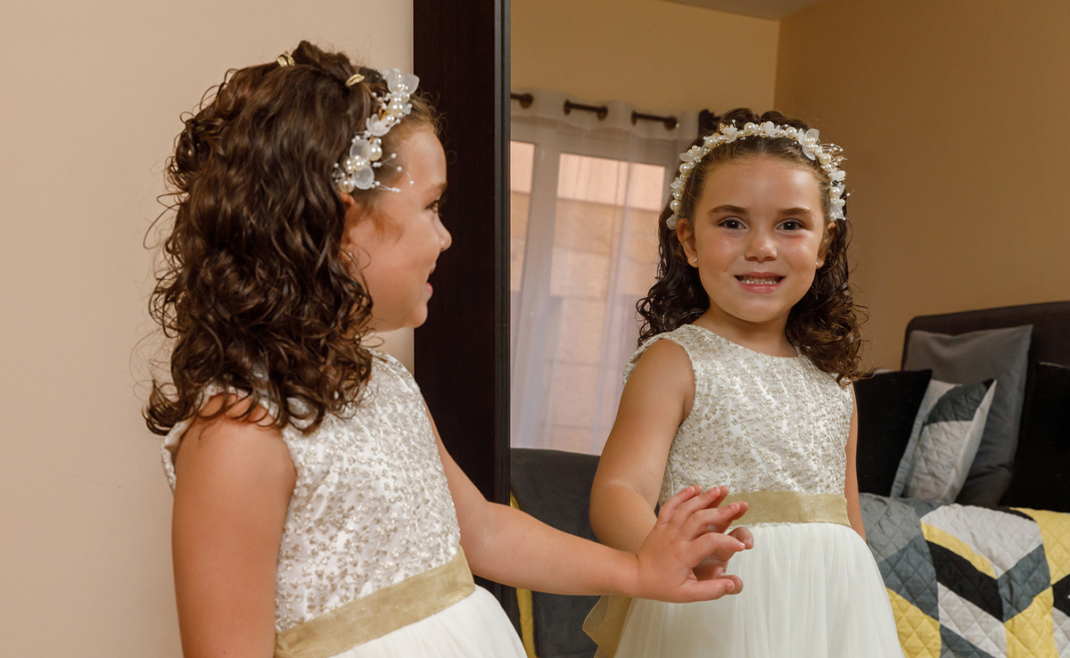 Com um sorriso contagiante, Iris contempla o espelho, tocando-o suavemente, como quem se descobre num dia especial.