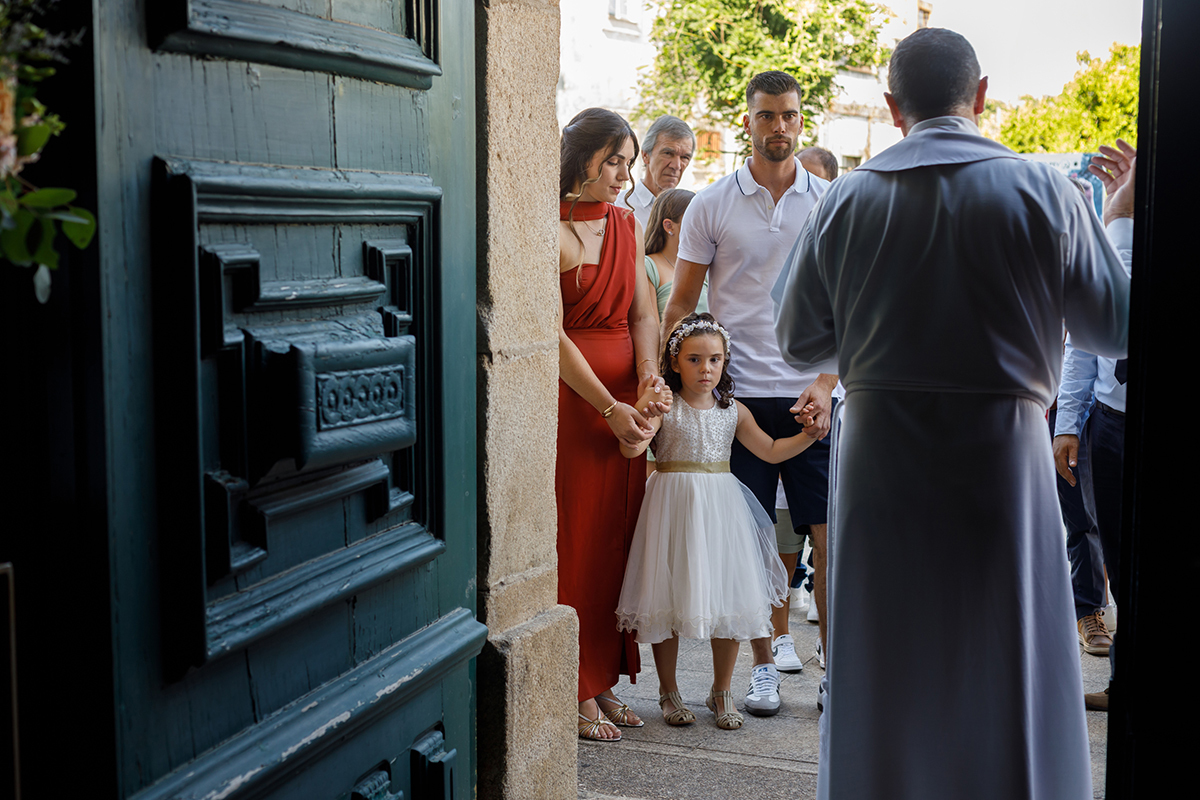 De mãos dadas com os pais, Iris prepara-se para entrar na igreja, num momento solene observado pelo padre e pelos convidados, um instante cheio de significado numa sessão de batizado.