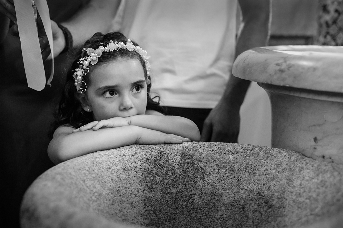 Em preto e branco, Iris descansa o rosto sobre os braços, pousados na Pia Batismal, com um olhar sereno e pensativo.