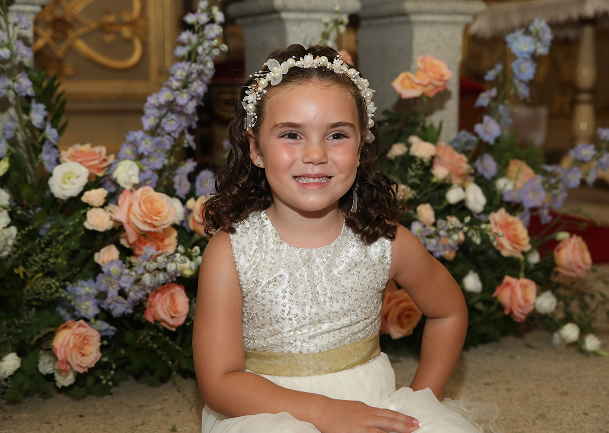 Com um sorriso radiante, Iris está sentada na igreja, rodeada pela beleza dos arranjos florais.