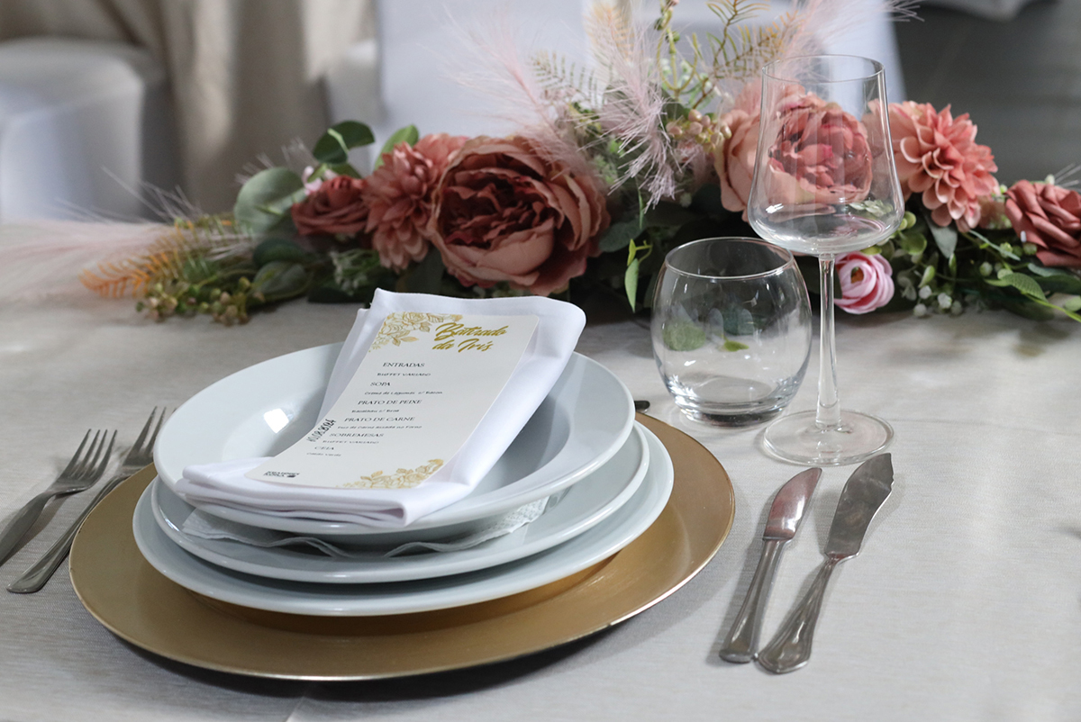 Promenor decoraçao , talheres, copos e um papel com a ementa compõem a mesa, onde flores dão um toque especial.