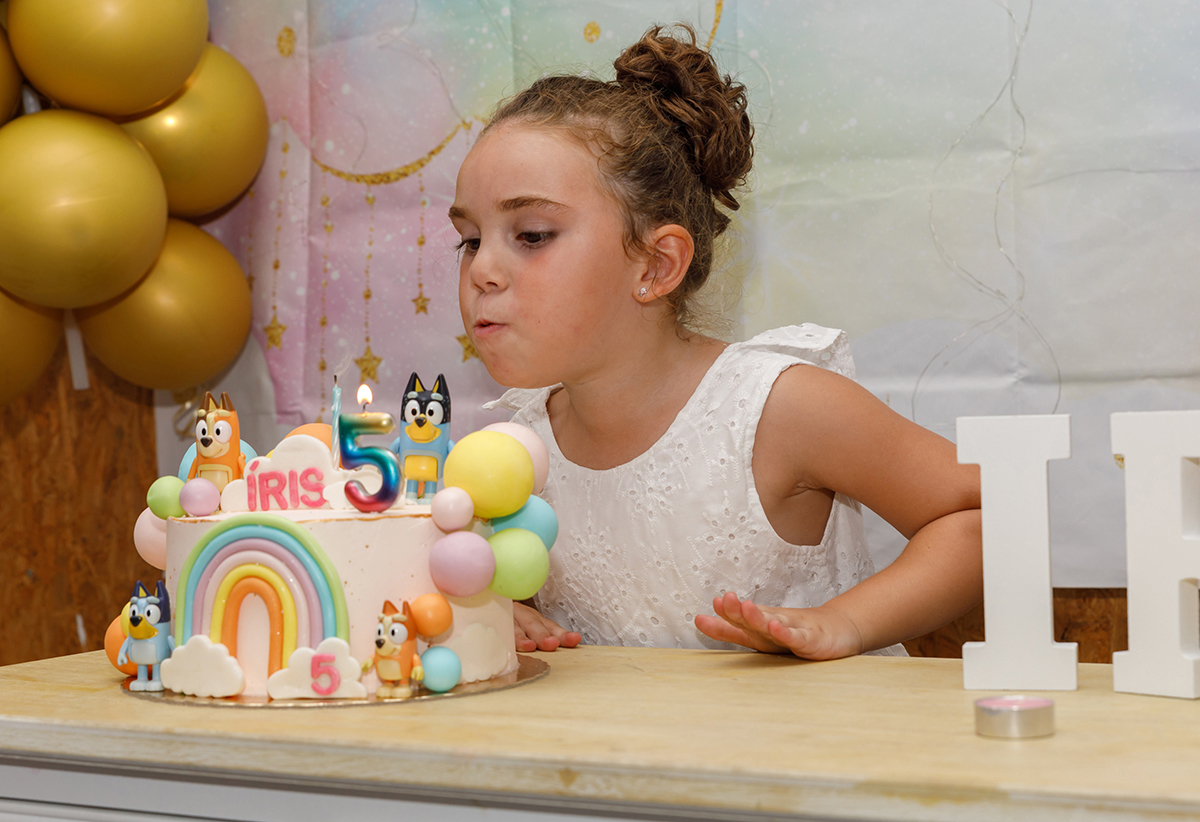 Iris, a celebrar os seus 5 anos, sopra a vela do bolo num cenário encantador em tons de rosa e verde, rodeada de estrelinhas, balões dourados.
