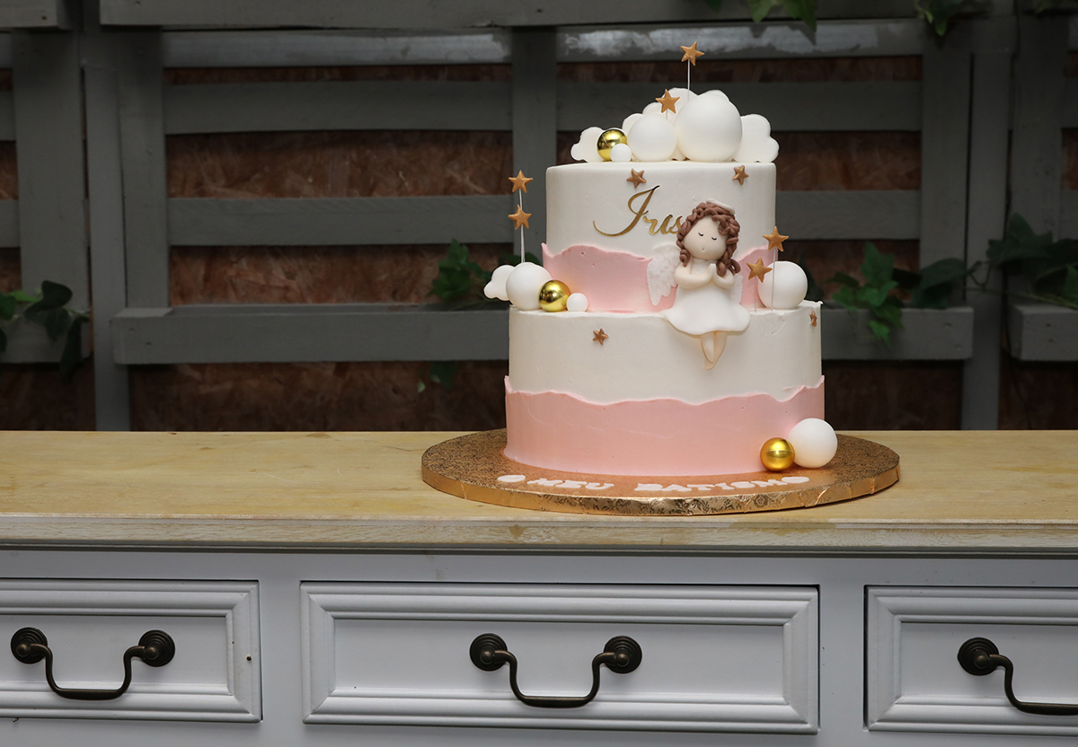 O bolo de batizado da Iris, em tons de branco e rosa, com duas camadas e uma doce menina sentada no andar de baixo, adornado com bolinhas douradas.