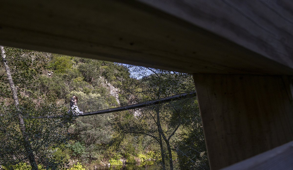 Entre o buraco do corrimão, um vislumbre precioso: ao fundo, Daniela e Jorge estão abraçados e a beijarem-se no meio da ponte, captando um momento íntimo através de uma moldura natural.