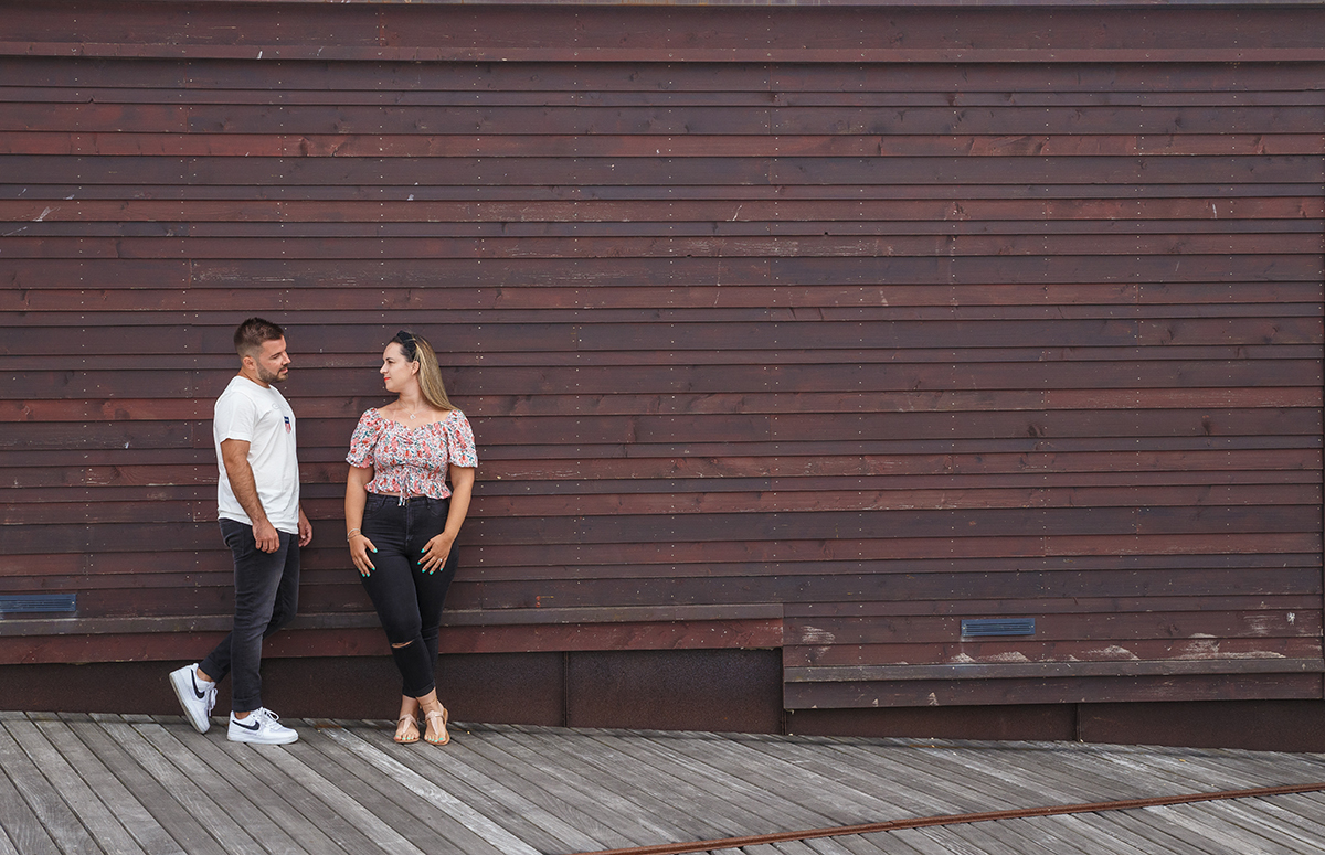 Encostados a uma parede de madeira, os dois estão juntos no lado esquerdo da imagem, Ricardo de perfil contempla Marta com ternura, enquanto ela olha para ele com o rosto voltado de lado e um sorriso subtil.