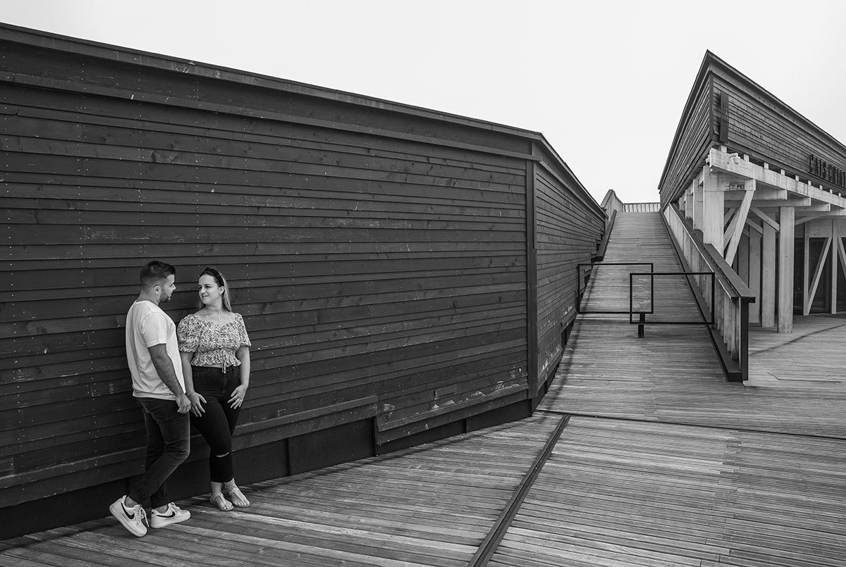 Em preto e branco, vemos novamente os dois encostados à parede de madeira, agora apanhados de outro ângulo onde os passadiços entram em cena, reforçando este cenário calmo onde os corpos conversam mais do que as vozes.