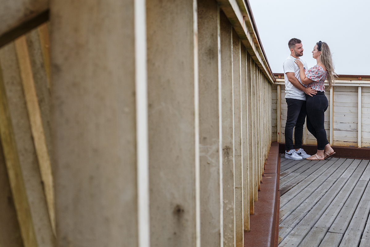 Encostada à varanda de madeira, a câmara observa discretamente os dois ao fundo: de perfil um para o outro, com Ricardo a segurar Marta pela cintura enquanto ela pousa as mãos no seu peito, e no ar sente-se uma serenidade que aquece.