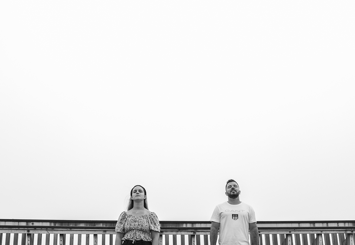 Em preto e branco, os dois estão encostados ao corrimão com um espaço simbólico entre eles, ambos com os olhos fechados e os rostos erguidos, como quem respira fundo e reflete juntos, num silêncio que une mais do que mil palavras.