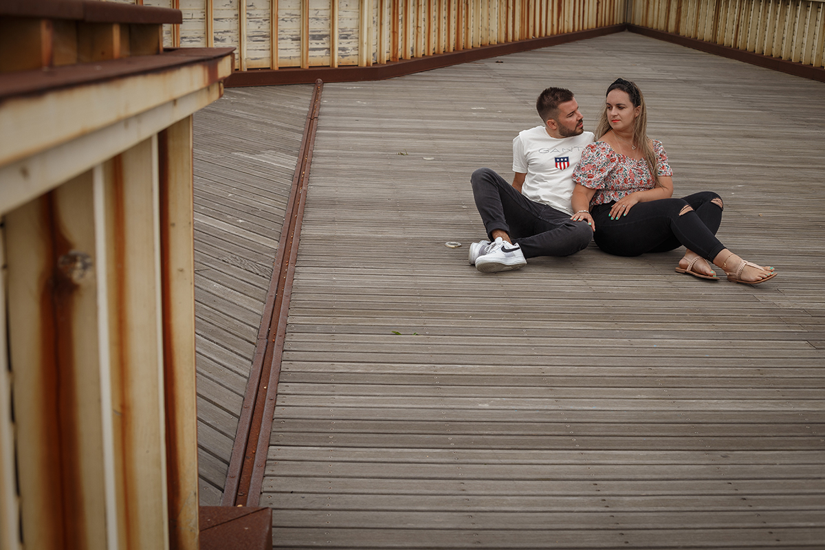 Sentados nos passadiços de madeira, Ricardo repousa com as mãos no chão atrás de si, enquanto Marta, ligeiramente inclinada, pousa uma mão na sua perna e outra na dela, e os dois trocam olhares sérios mas doces, como quem reconhece o futuro nos olhos.