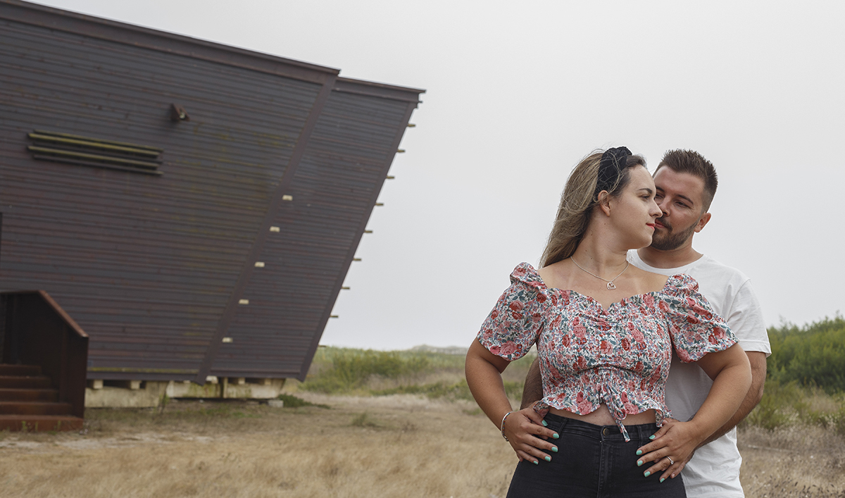 Do lado direito da imagem e da cintura para cima, Ricardo está atrás e envolve Marta com as mãos na cintura, enquanto ela retribui o gesto colocando as suas mãos sobre as dele, e o olhar dela segue em frente enquanto ele a contempla com ternura silenciosa