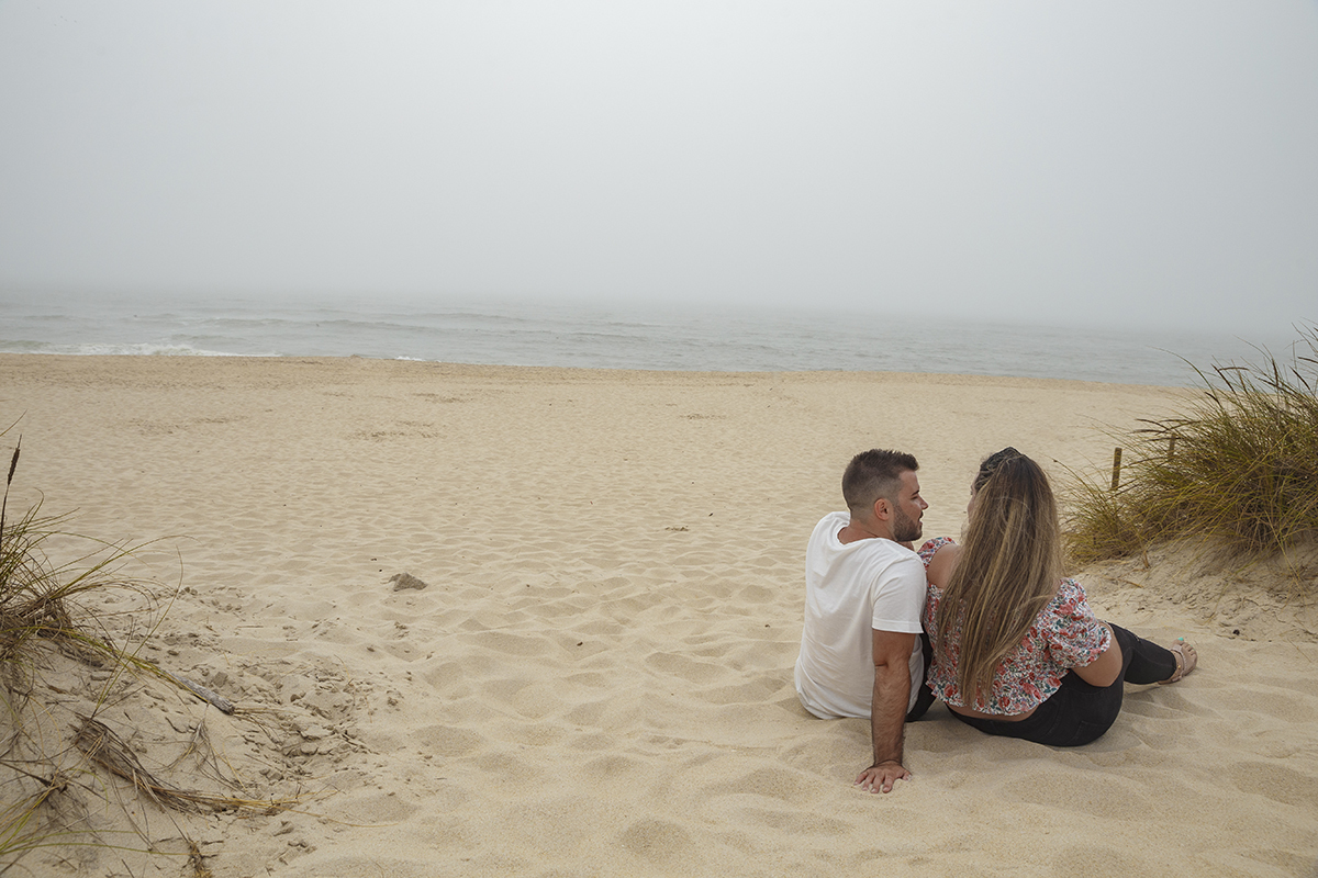 Aqui vemos os dois de costas, deitados na areia do lado direito da foto, com Marta a olhar para o mar em contemplação e Ricardo a olhar para ela com um leve sorriso, num instante de silêncio cúmplice.