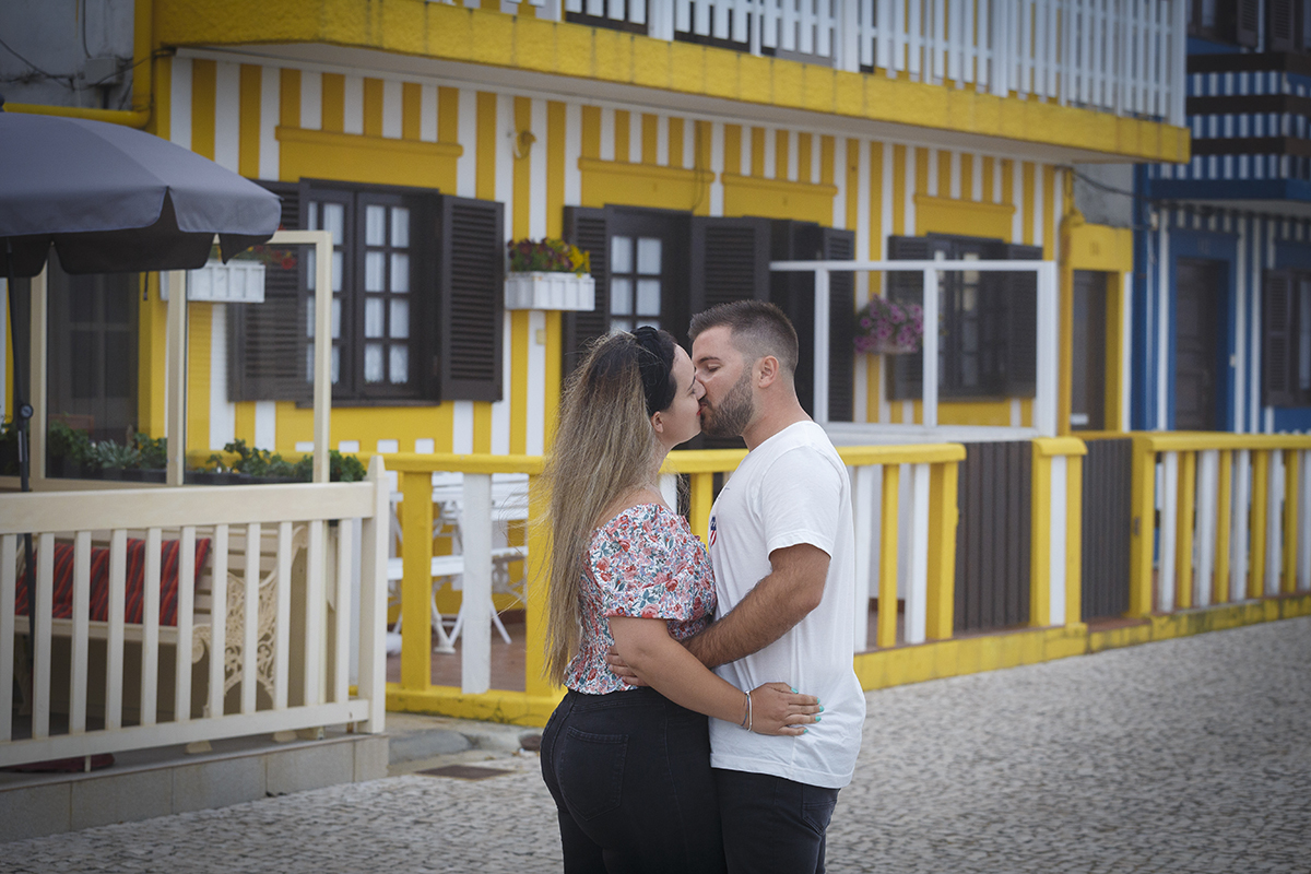 Aqui vemos as casinhas coloridas da Praia da Costa Nova, e ao fundo uma amarela que dá um toque alegre ao cenário, vemos os dois da coxa para cima, frente a frente a abraçarem-se pela cintura e a beijarem-se com ternura.