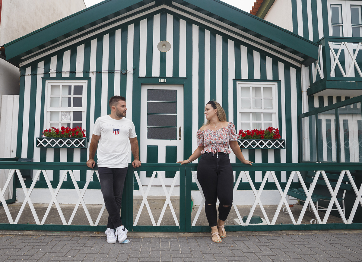 Aqui com o cenário de uma casinha verde e branca, os dois estão encostados ao varão e apoiam-se com as mãos no corrimão, com um espaço entre eles, a olharem-se felizes e sorridentes como se cada risinho fosse uma promessa leve.