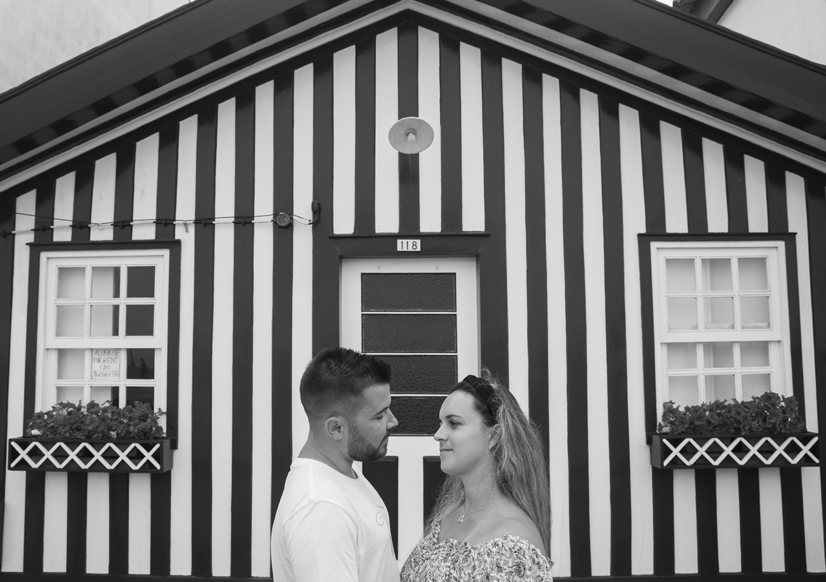 Aqui temos uma foto a preto e branco, onde os vemos do peito para cima frente a frente a encararem-se com felicidade, e atrás uma casinha que faz pano de fundo a este instante doce.