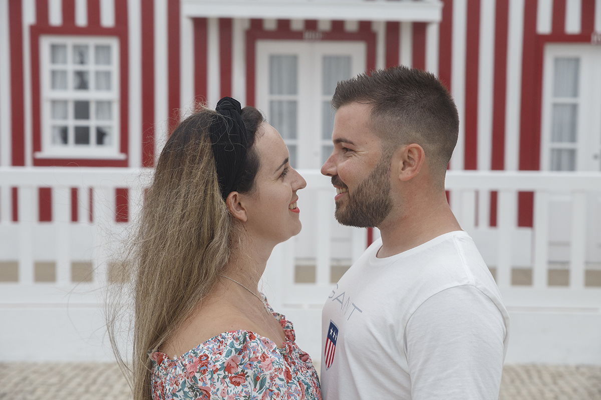 Aqui vemos um plano mais aproximado, do peito para cima, com os dois a sorrirem um para o outro num olhar cheio de carinho, e atrás o fundo desfocado deixa entrever uma casinha vermelha e branca.