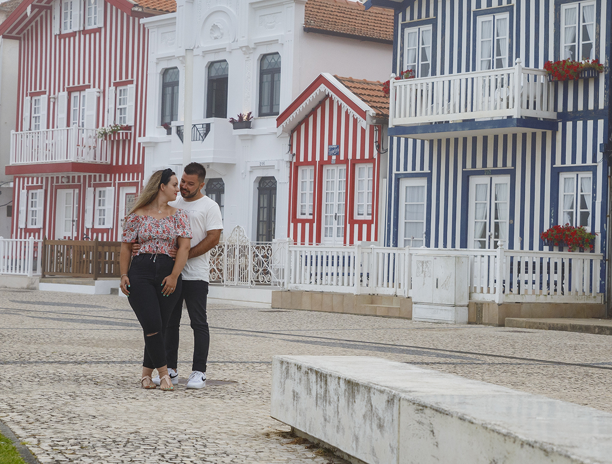 Aqui vemos os dois num plano mais afastado, com casinhas coloridas ao fundo, brancas, vermelhas e azuis, e do lado esquerdo da imagem estão lado a lado, com Ricardo ligeiramente atrás a segurar a cintura de Marta, que olha para trás com um sorriso doce.