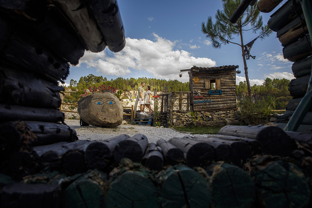 Dentro de um círculo mágico de troncos entrelaçados, os dois encontram-se na base das escadas de uma pequena casa rústica com pedra à vista, frente a frente como se fossem personagens de um conto encantado.