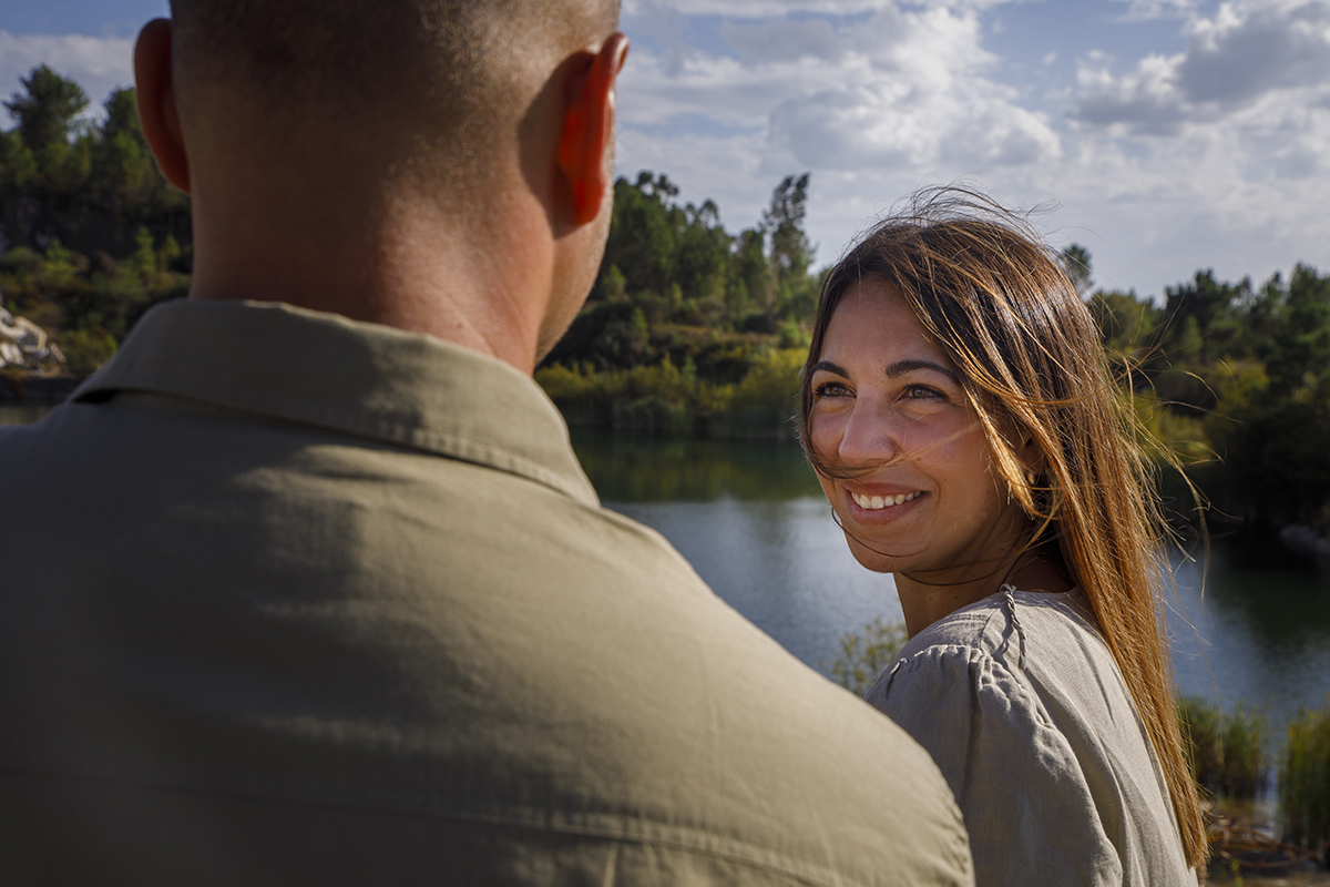 Em plano aproximado dos ombros para cima, vemos Bruno de costas, desfocado, enquanto Patrícia aparece à frente, com o rosto virado para trás. Ela olha para ele com um sorriso encantador, meio tímido meio cúmplice, e a imagem parece suspensa no tempo.