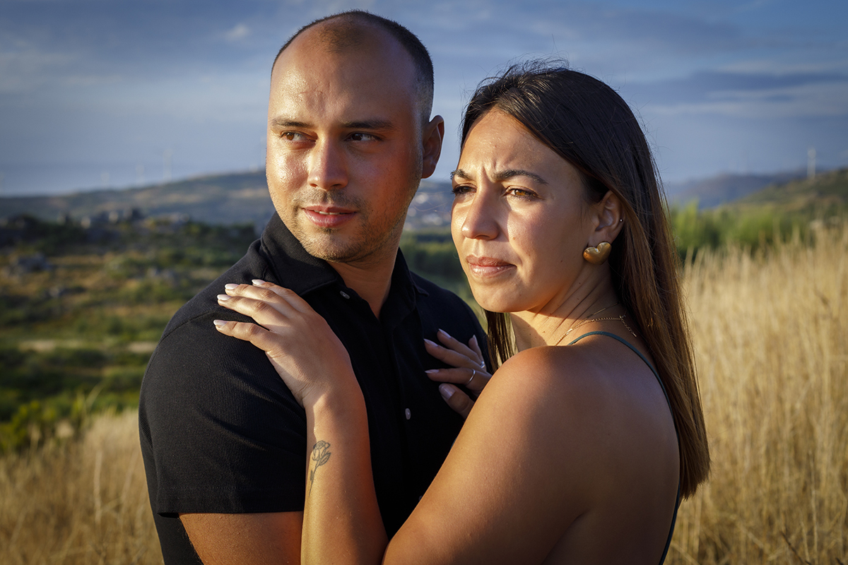 Num plano mais próximo, do peito para cima, os dois estão frente a frente, Patrícia com uma mão no ombro e outra no peito de Bruno, ambos com olhares sérios, mas repletos de felicidade e pensamentos profundos, enquanto o sol os aquece suavemente.