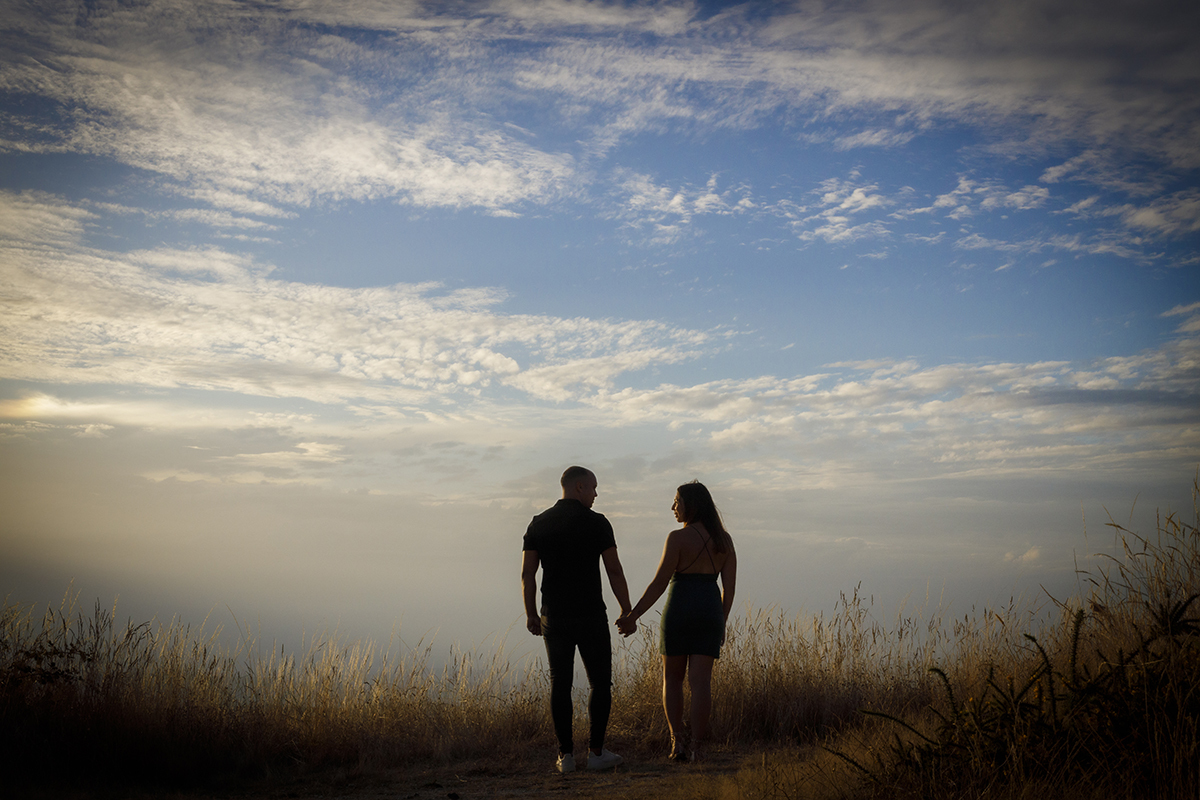 Uma vinheta envolve a imagem central onde os dois aparecem de costas para a câmara, mãos dadas, de pé num campo castanho sob um céu azul com leve nevoeiro, olhando um para o outro com um amor silencioso e profundo.