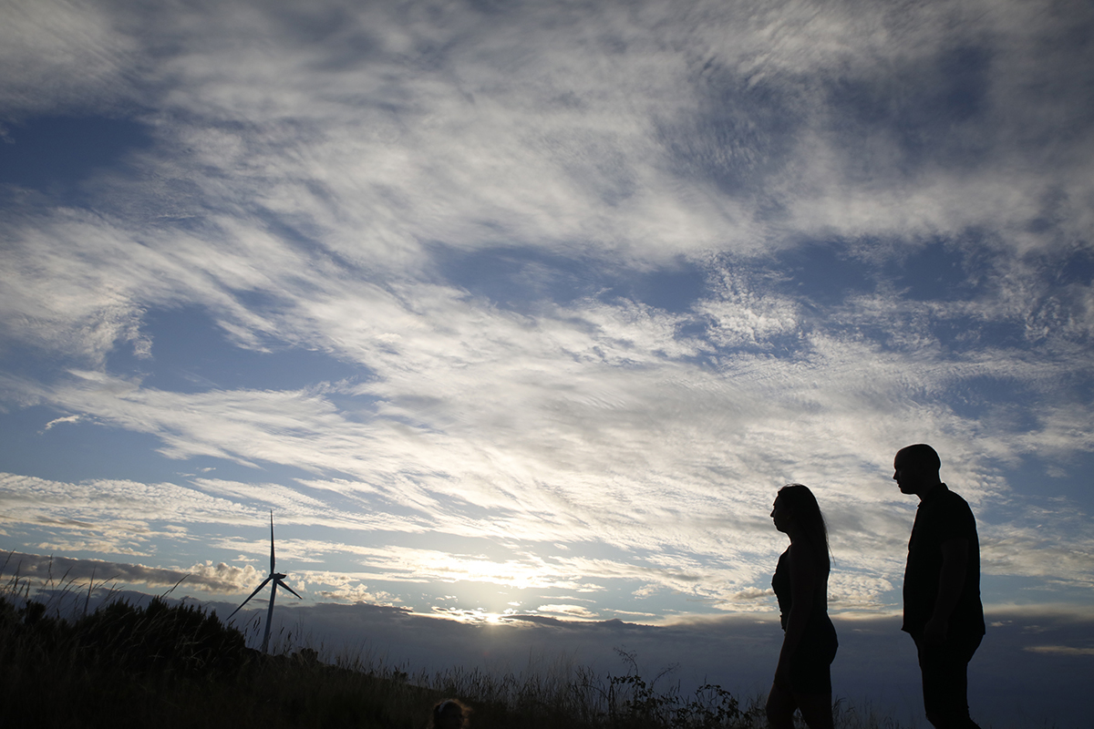 Num plano mais afastado, os dois aparecem no lado direito da imagem, ainda em silhueta, sob um céu azul limpo com nuvens leves e uma ventoinha eólica que se ergue ao fundo, sugerindo o vento suave que acompanha o amor deles.