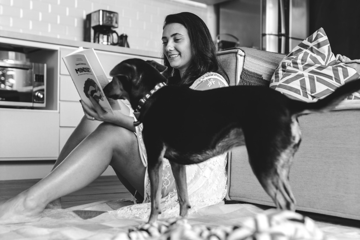 foto em preto e branco de mulher sentada no chão da sala lendo livro enquanto cachorra passa na frente