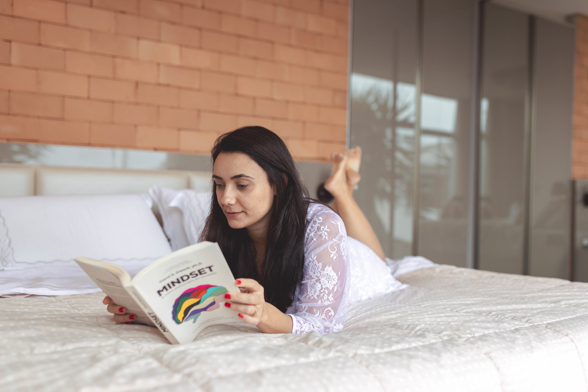 foto de mulher de camisola e hobby brancos, deitada de bruços na cama lendo livro