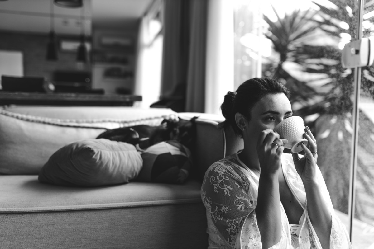 foto em preto e branco de mulher sentada no chão da sala, encostada no sofá tomando uma xícara de chá, enquanto o cachorro está deitado no sofá.