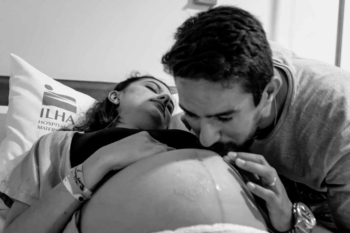 homem dando beijo na barriga da mulher grávida deitada na cama do quarto da maternidade