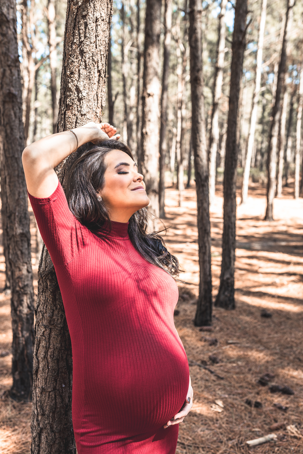 gestante de vestido vermelho, com os olhos fechados encostada em árvore de parque em Florianópolis