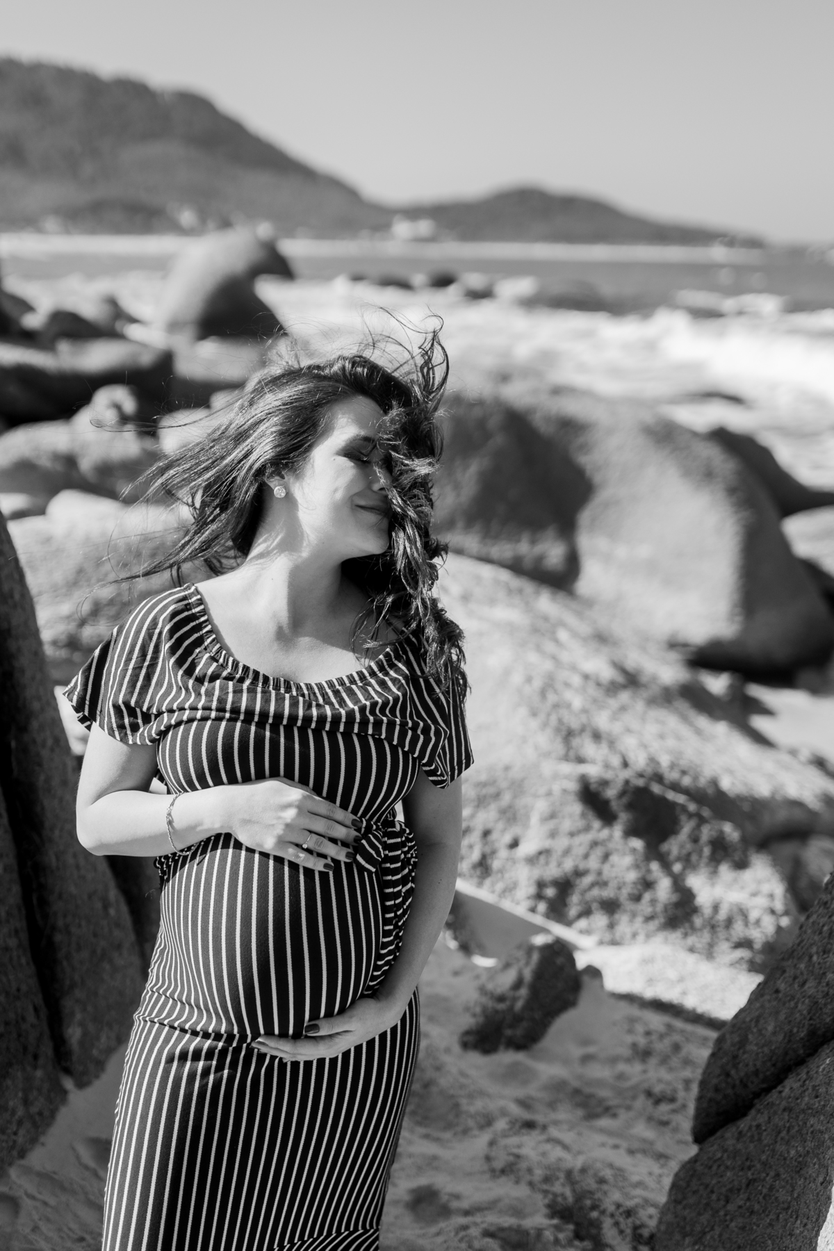 gestante de vestido na praia com os cabelos no rosto. foto em preto e branco