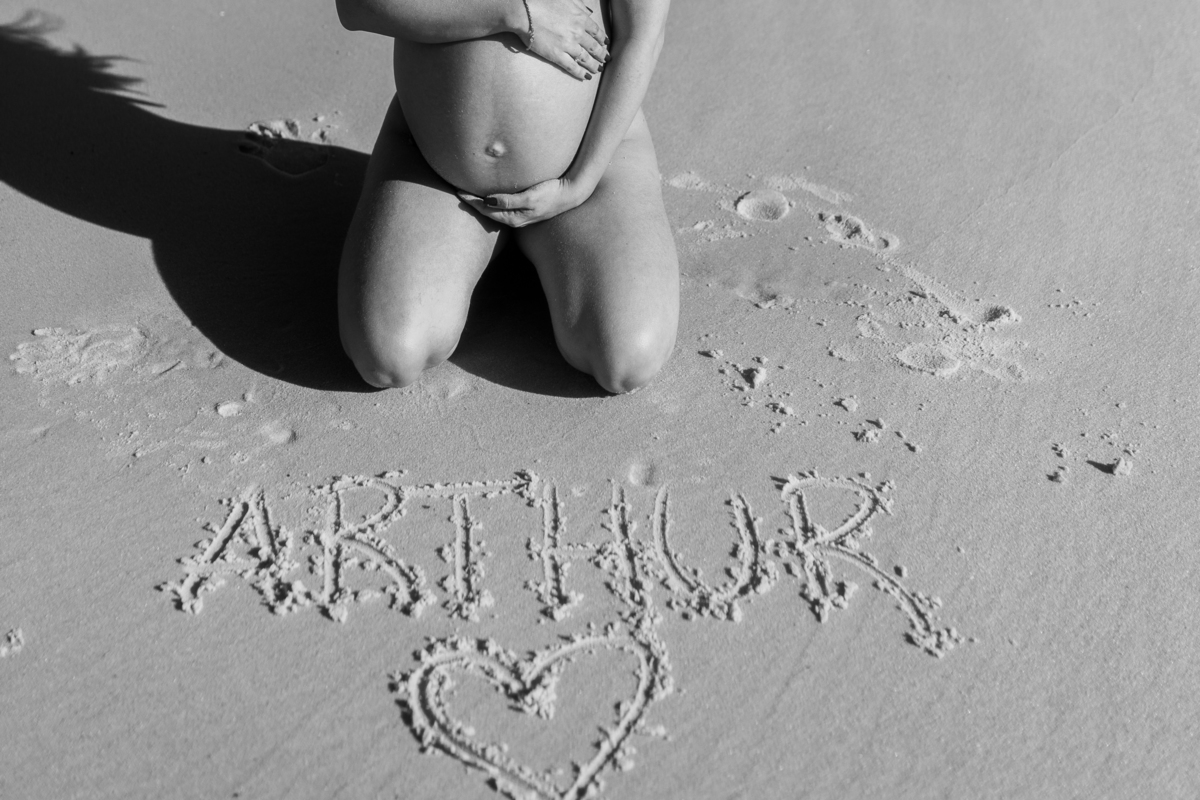 Gestante ajoelhada na areia envolvendo a barriga com o nome do seu filho 