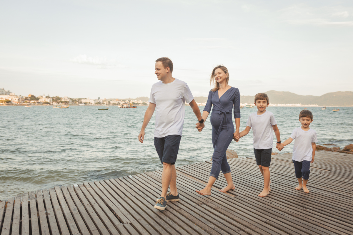 pai, mãe grávida e os dois filhos de mãos dadas andando no trapiche da praia em Florianópolis