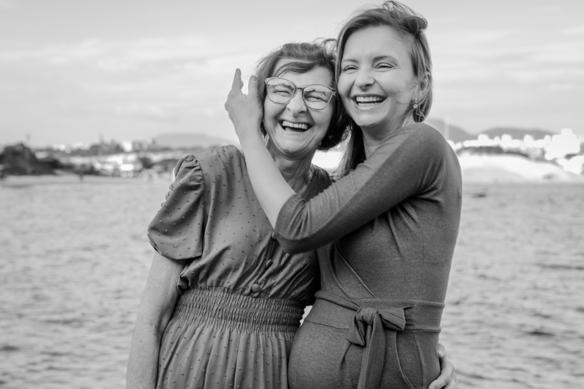 mã e efilha grávida sorrindo abraçadas na praia. Foto em preto e branco