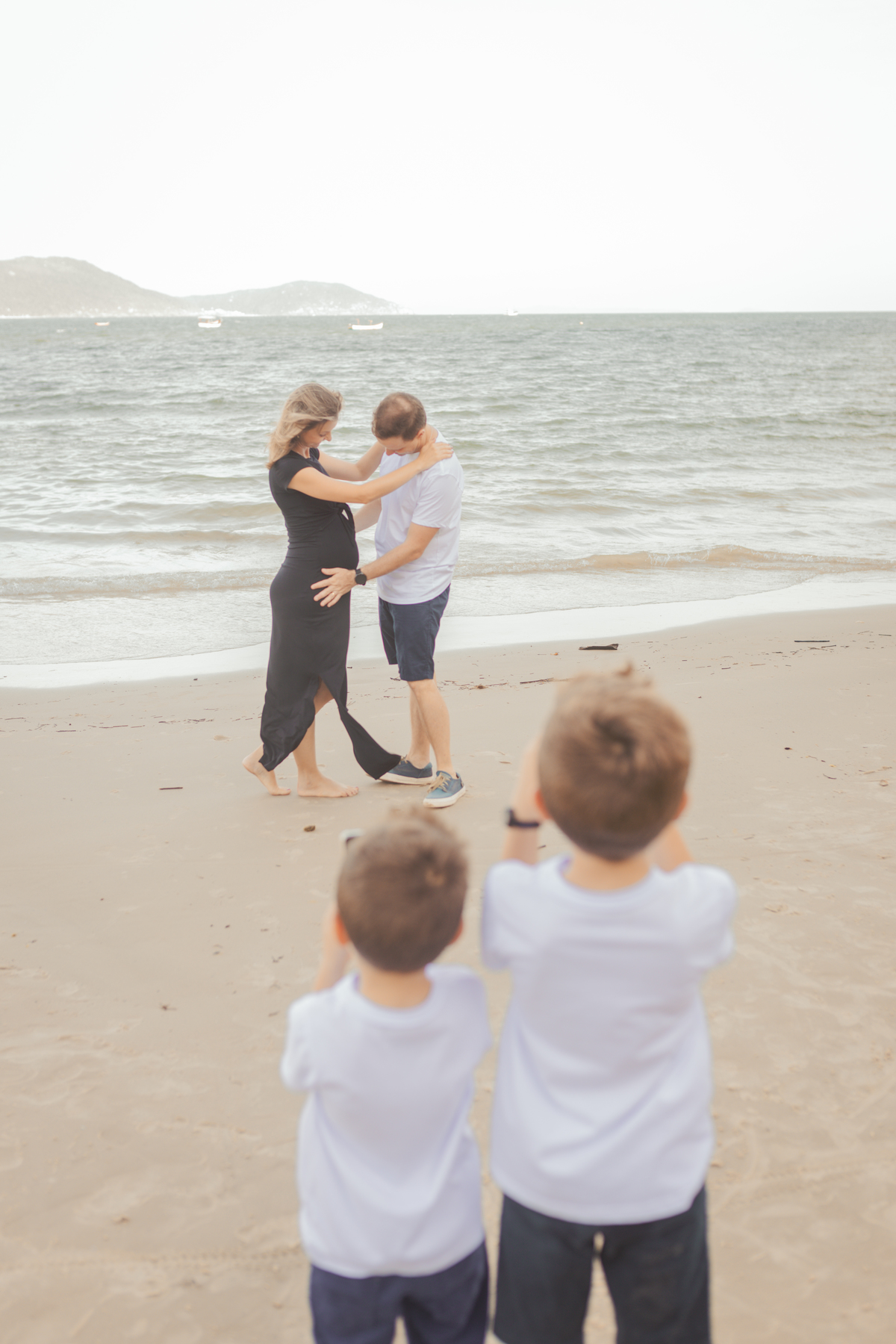 filhos tirando foto dos pais, enquanto o pai olha a barriga da mãe grávida na praia em Florianópolis