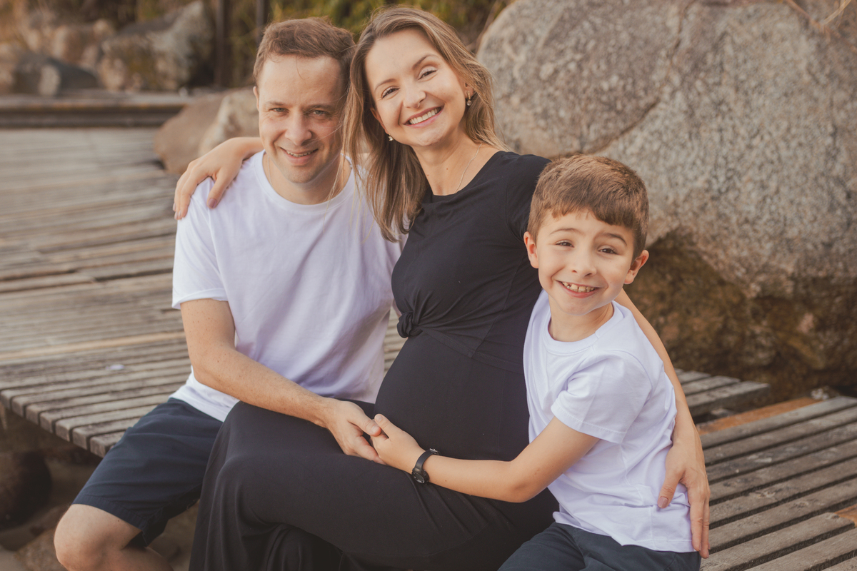 Marido e filho abraçados na mãe grávida. Sentados no trapiche da praia.
