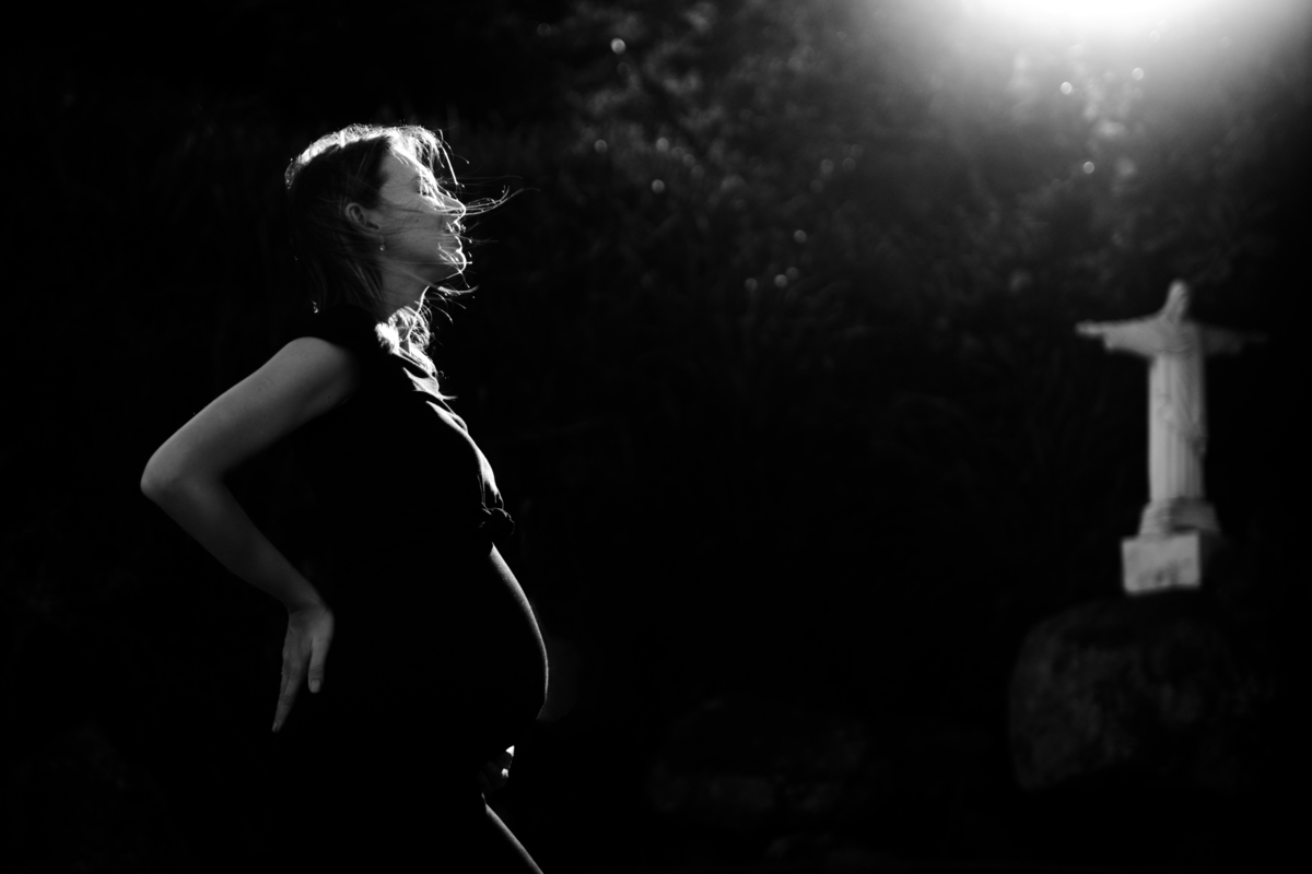Gestante com a mão na barriga no nascer do sol próxima a imagem do cristo. foto em preto e branco