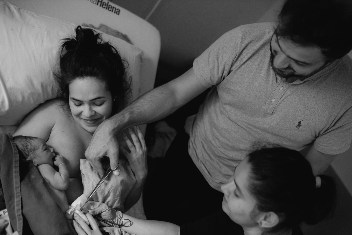 foto em preto e branco de bebê no colo da mãe que está sentada na cama, enquanto o pai corta o cordão umbilical com o auxílio da enfermeira.