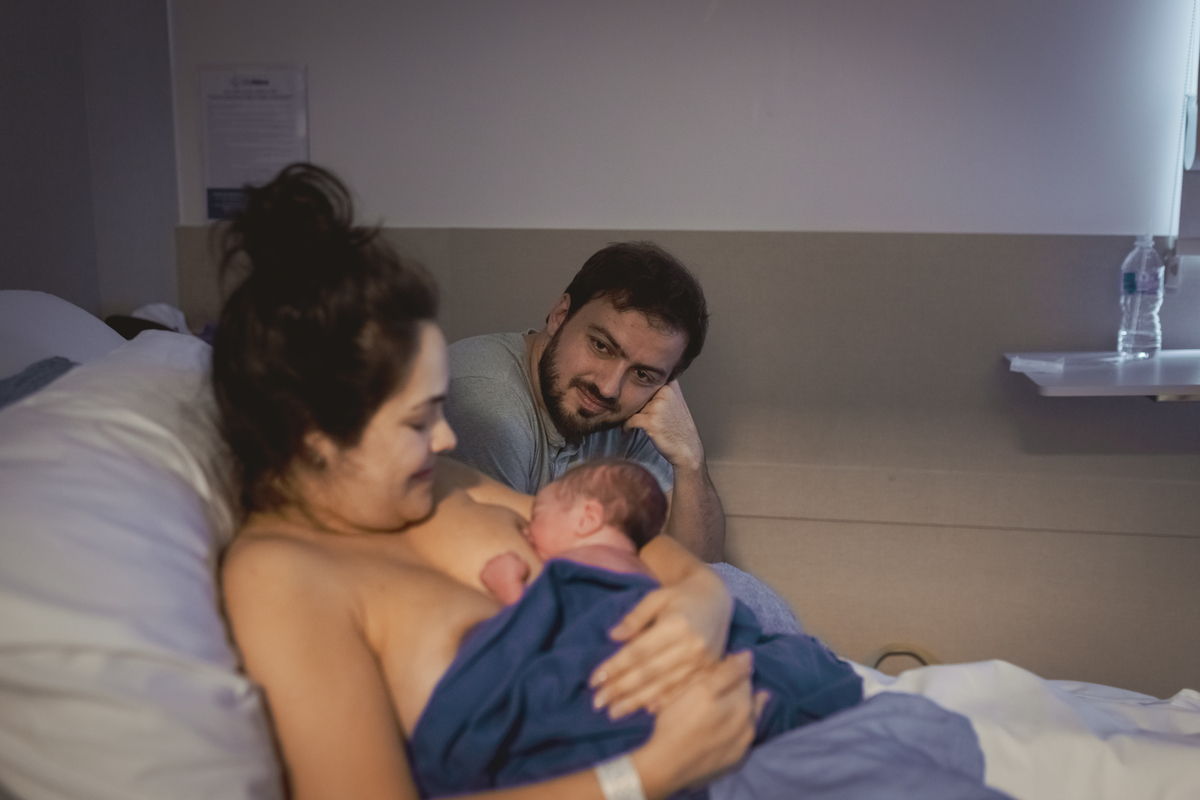 mãe amamentando bebê logo após o nascimento enquanto marido está sentado na cadeira de acompanhante admirando.