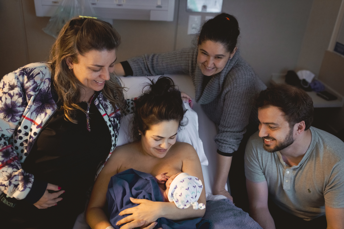 mãe sentada na cama da sala de parto carregando o bebê enquanto pai e equipe médica admiram ele.