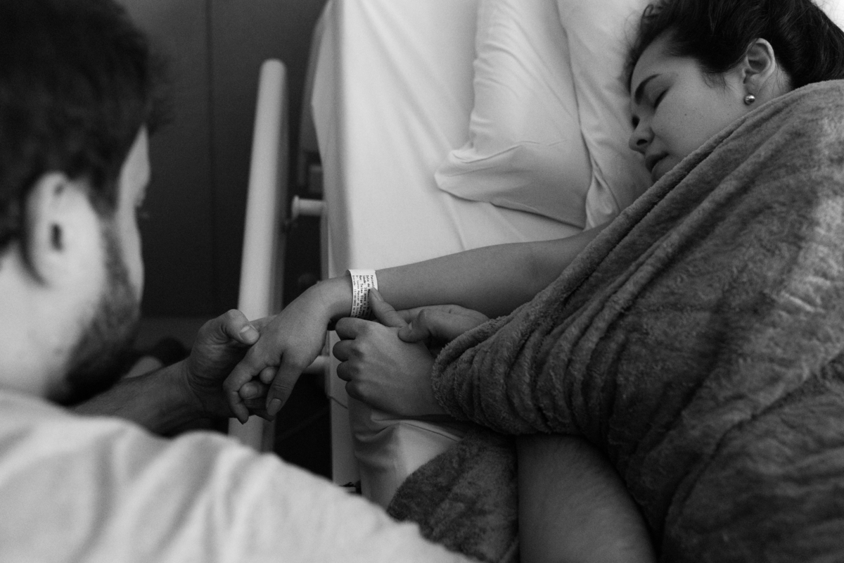 foto em preto e branco de marido segurando as mãos de gestante que está deitada na cama em trabalho de parto