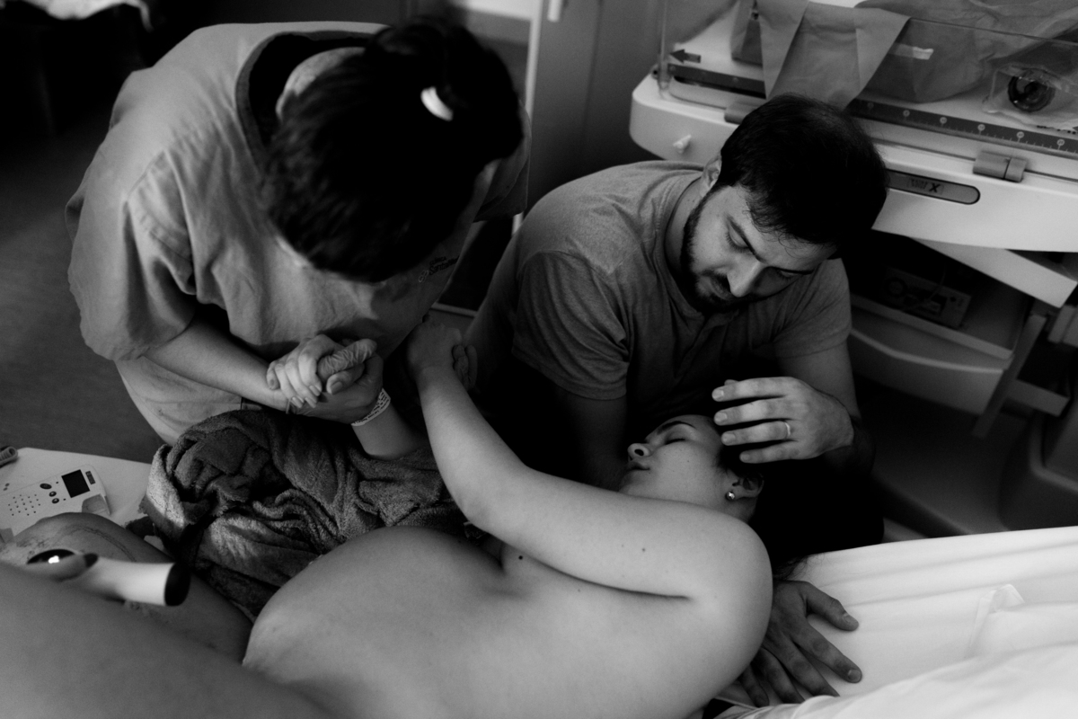 foto em preto e branco de gestante deitada na cama da sala de parto recebendo suporte do marido e da enfermeira