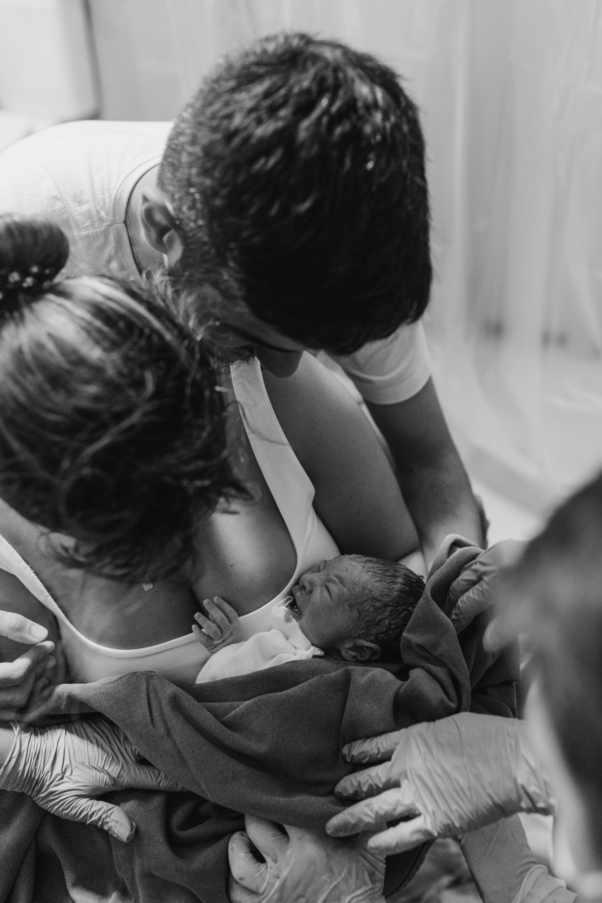 foto em preto e branco de bebê no colo da mãe logo após o nascimento, e pai atrás olhando para o bebê.