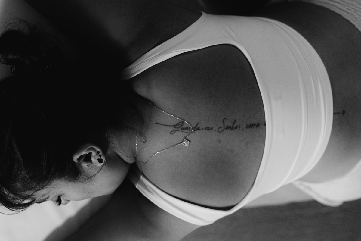 foto em preto e branco feita de cima, de gestante com tatuagem nas costas, apoiada na beira da cama.