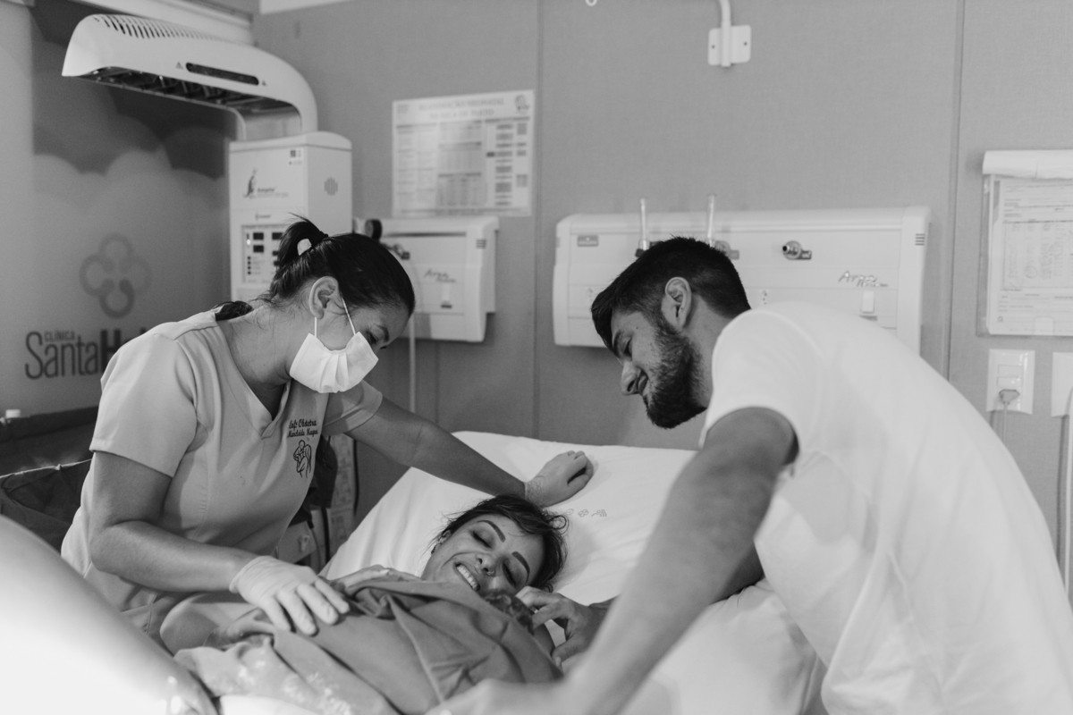 foto em preto e branco de mãe deitada no leito da sala de parto carregando seu filho , doula e marido, todos olhando para o bebê