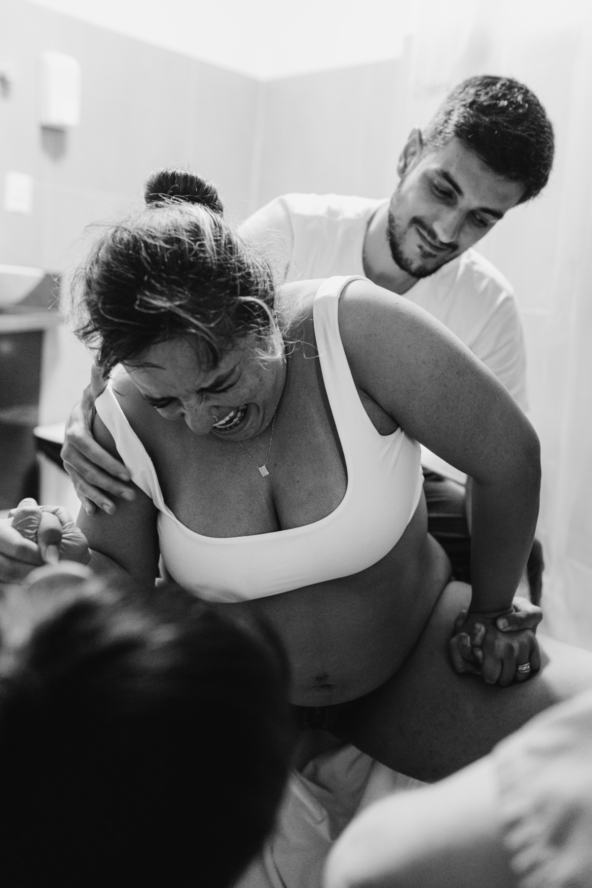 foto em preto e branco de gestante sentada em banquinho, tendo uma contração, e marido atrás segurando ela