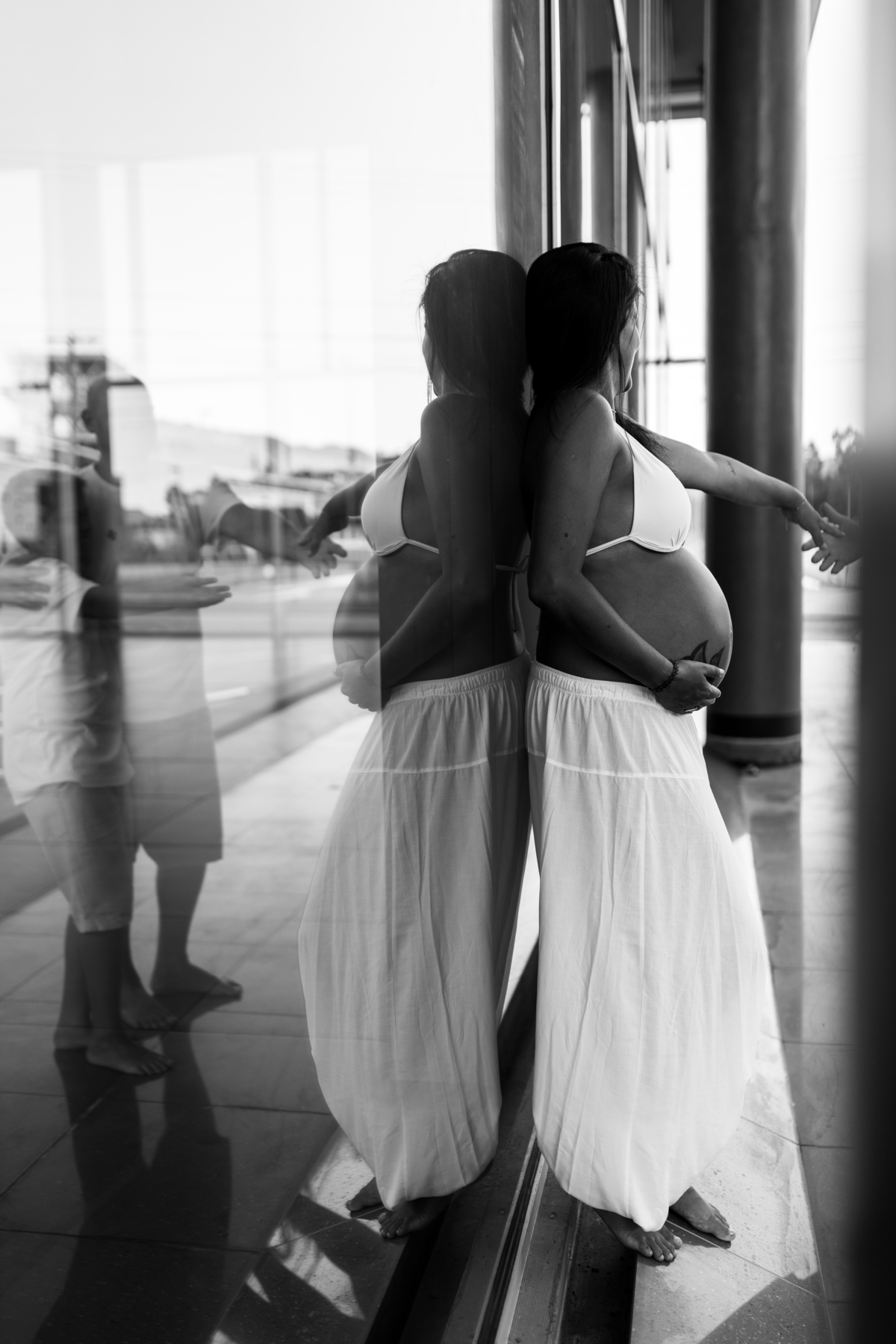 gestante encostada em vidro dando a mao para o marido e o filho que aparecem no reflexo do vidro