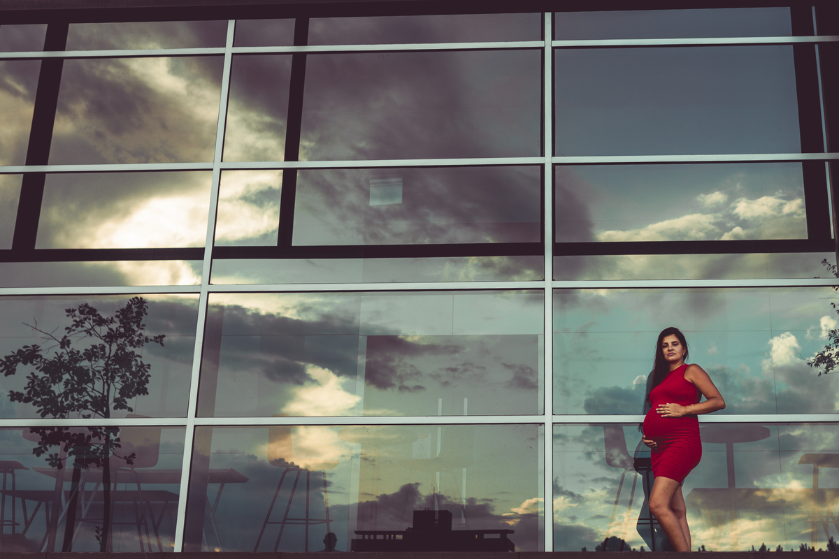 gestante de vestido vermelho encostada em vidro com reflexo da natureza