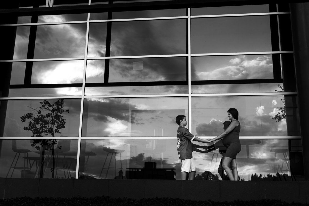 foto em preto e branco de mãe gestante e filho de mãos dadas em frente a vidro com reflexo da natureza