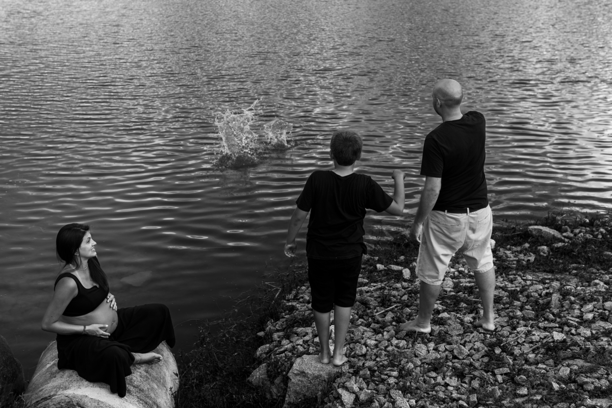 foto em preto e branco de gestante sentada olhando para o marido e o filho enquanto eles brincam de jogar pedra no lago